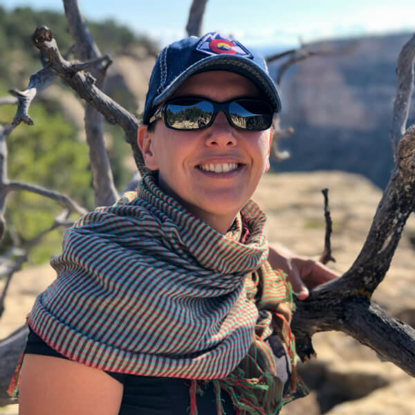 Delphine, the owner, posing for a photo in front of a bare tree with sunglasses and a hat on