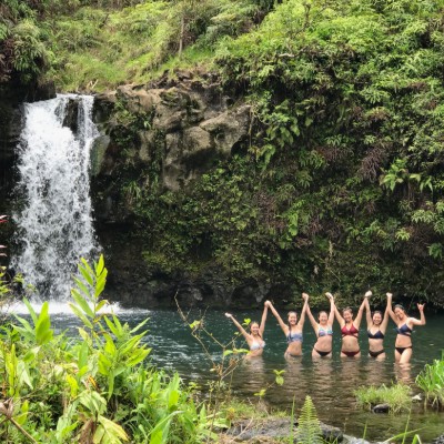 A group of girls in their swimming costumes swimming in a pool fed by a large waterfall